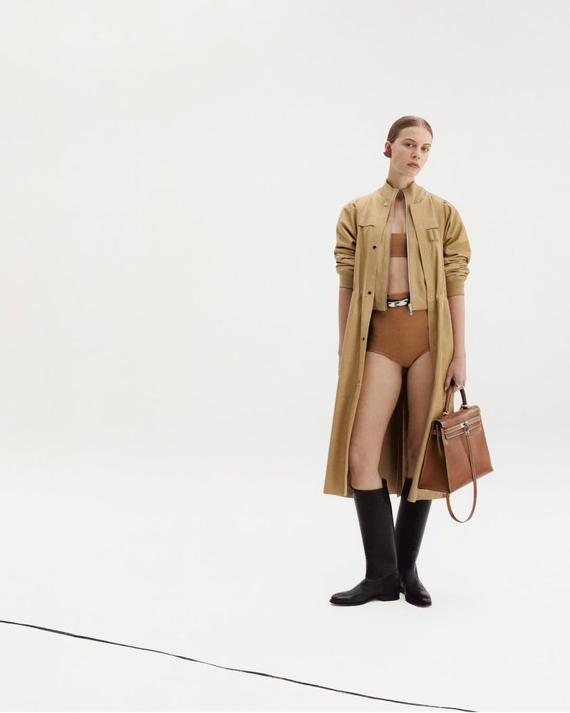 A woman in a trench coat, bikini, and boots poses with a brown handbag against a white backdrop, fashion forward and elegant.