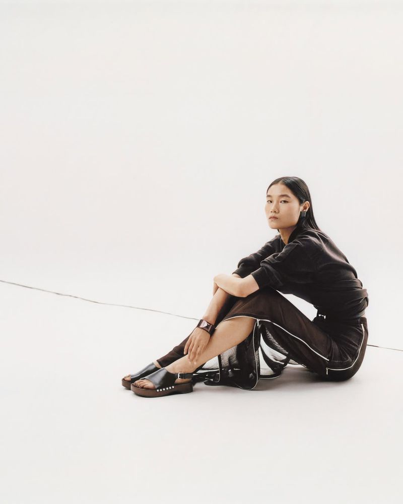 An Asian woman in a dark sweatshirt and skirt sits in a white studio wearing black studded sandals and a bracelet in a medium shot.