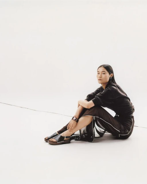 An Asian woman in a dark sweatshirt and skirt sits in a white studio wearing black studded sandals and a bracelet in a medium shot.