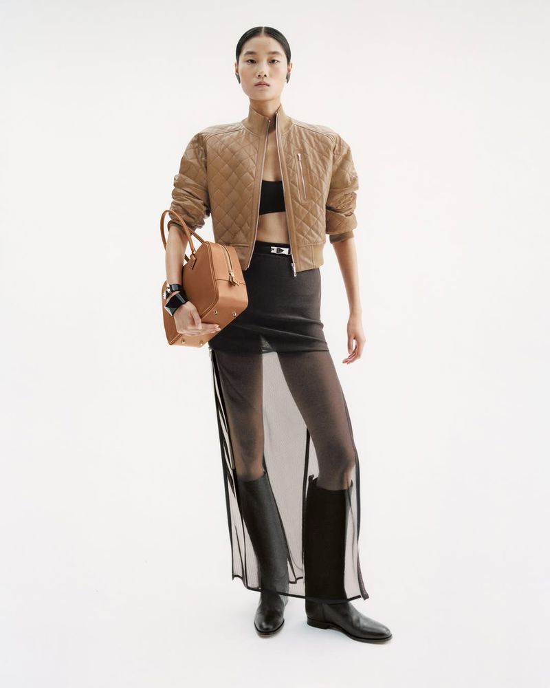 An Asian woman poses in a studio wearing a tan jacket, black bra top, semi-sheer skirt, black boots, and holding a tan satchel bag.