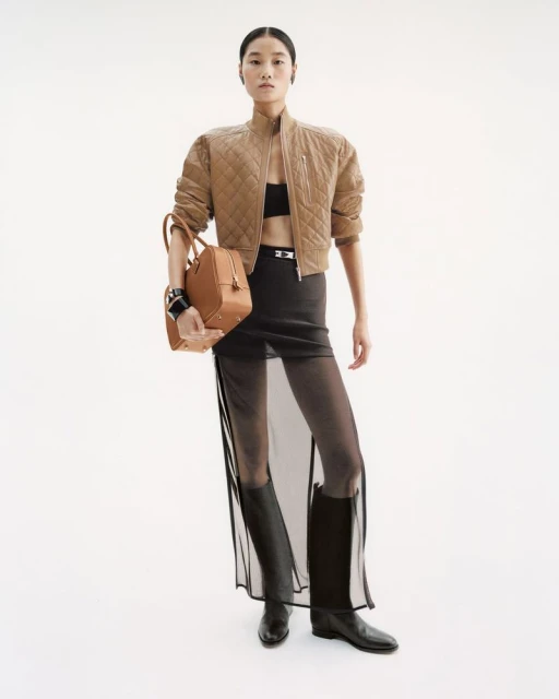 An Asian woman poses in a studio wearing a tan jacket, black bra top, semi-sheer skirt, black boots, and holding a tan satchel bag.