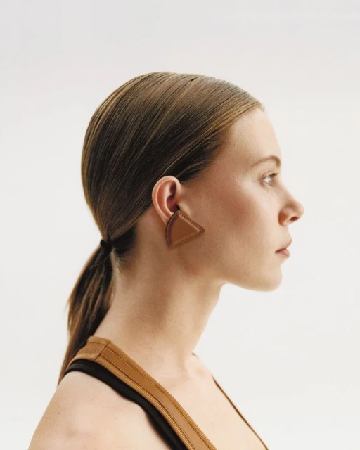 Profile shot of a light-skinned adult woman with long brown hair in a ponytail, wearing a brown triangle earring, against a white background.