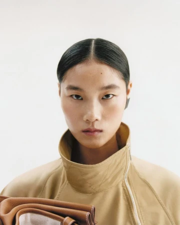 Portrait of an Asian female model in a tan zip-up jacket holding a brown and white purse against a white background.