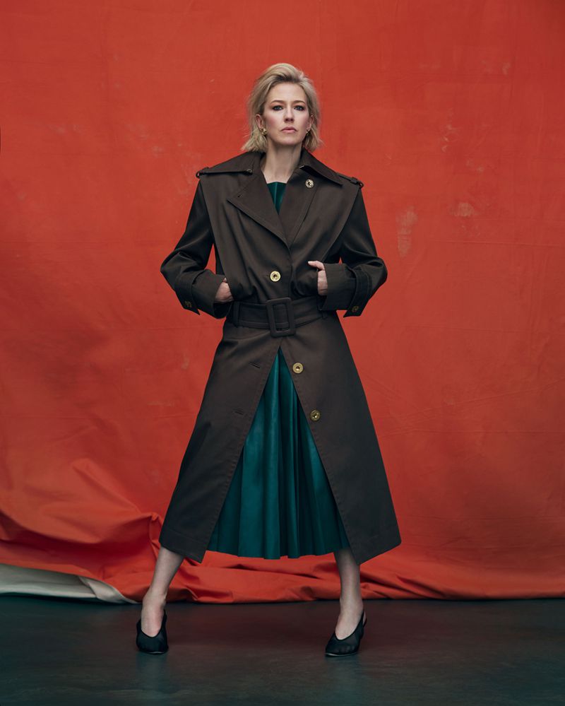 A full-length portrait showcases a blonde woman in a belted trench coat over a teal dress and black shoes against an orange backdrop.