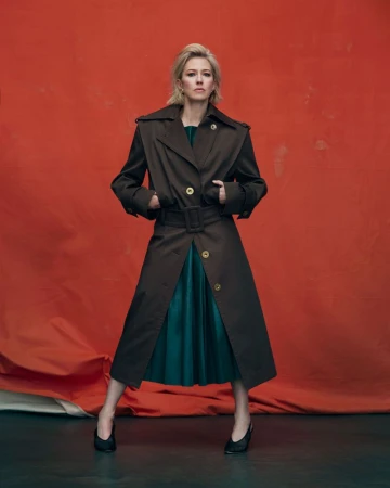A full-length portrait showcases a blonde woman in a belted trench coat over a teal dress and black shoes against an orange backdrop.