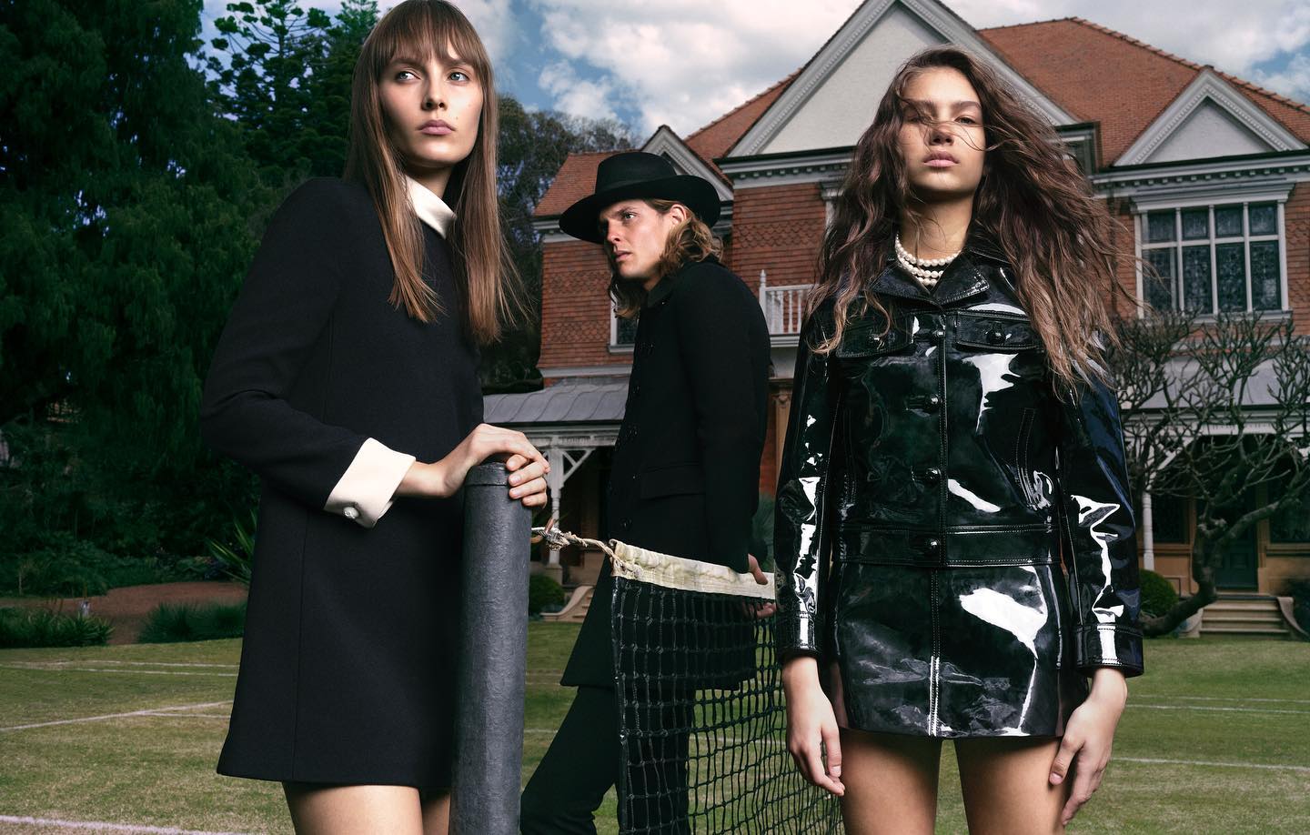 Three fashion models pose in front of a house on a lawn. Two women flank a man. One woman wears a black dress, the other a shiny black coat.