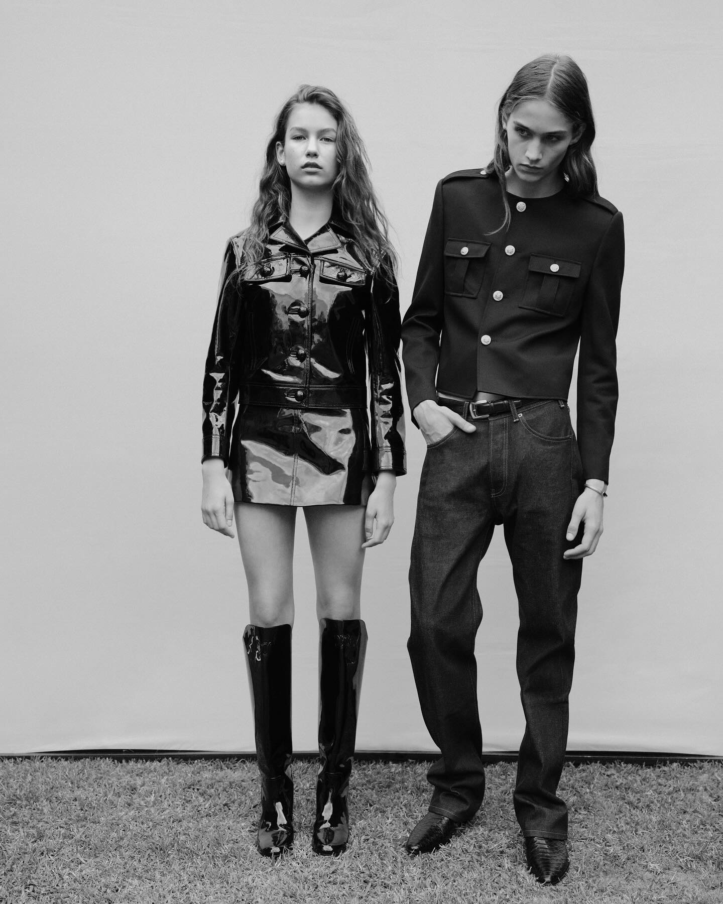 Black-and-white fashion shot of a young woman and man posing in glossy jackets, dark jeans, and boots on a patch of lawn against a blank backdrop.
