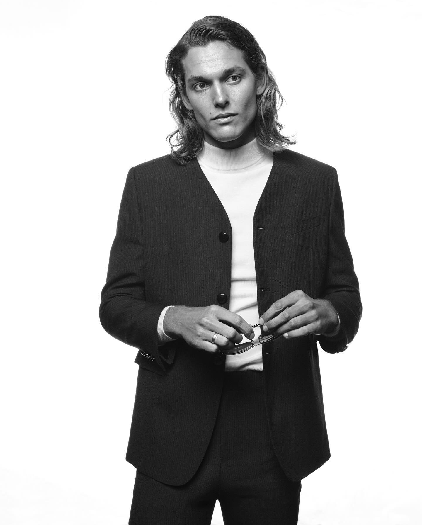 Man with long hair in dark collarless blazer holding sunglasses against a plain white background