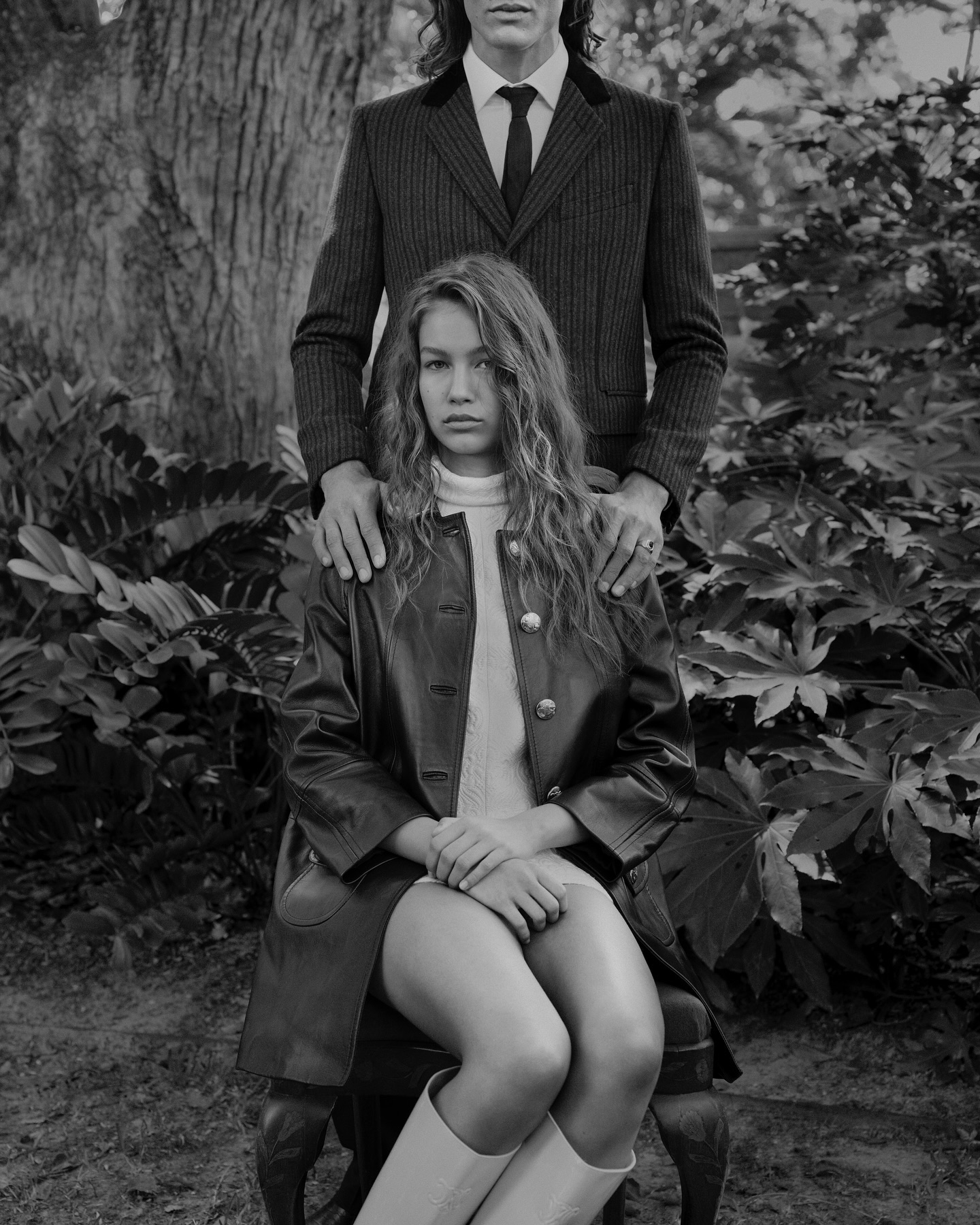 Monochrome fashion photo of a woman sitting on a chair in a dress and boots, with a man standing behind her in a suit.