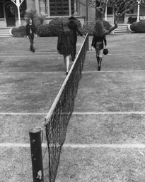 Black and white editorial image of multiple people walking on a lawn near a tennis net outside a building.