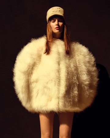Woman in an oversized, furry, white coat and matching hat posing against a dark background in a fashion editorial style.