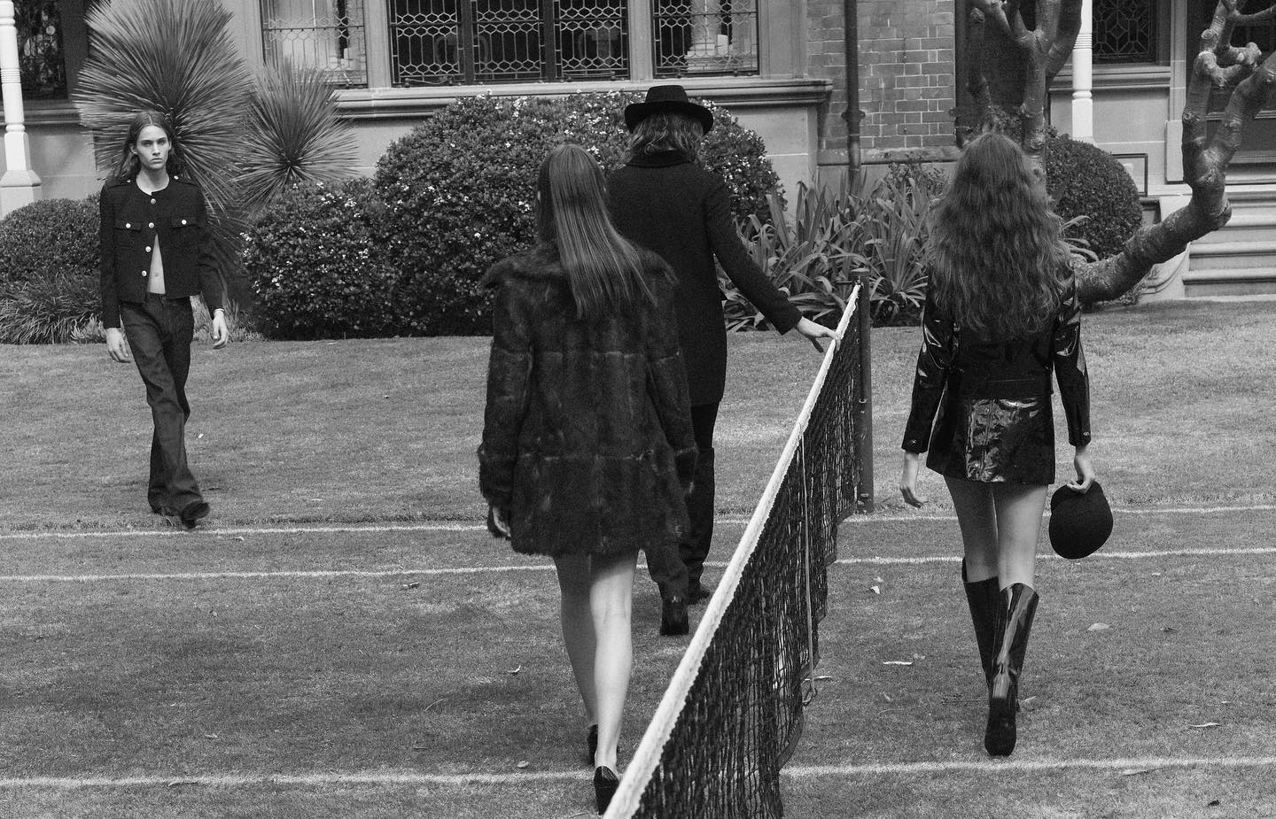 Black and white shot of four people walking on a tennis court, framed by the net, surrounded by grassy landscape and building backdrop.
