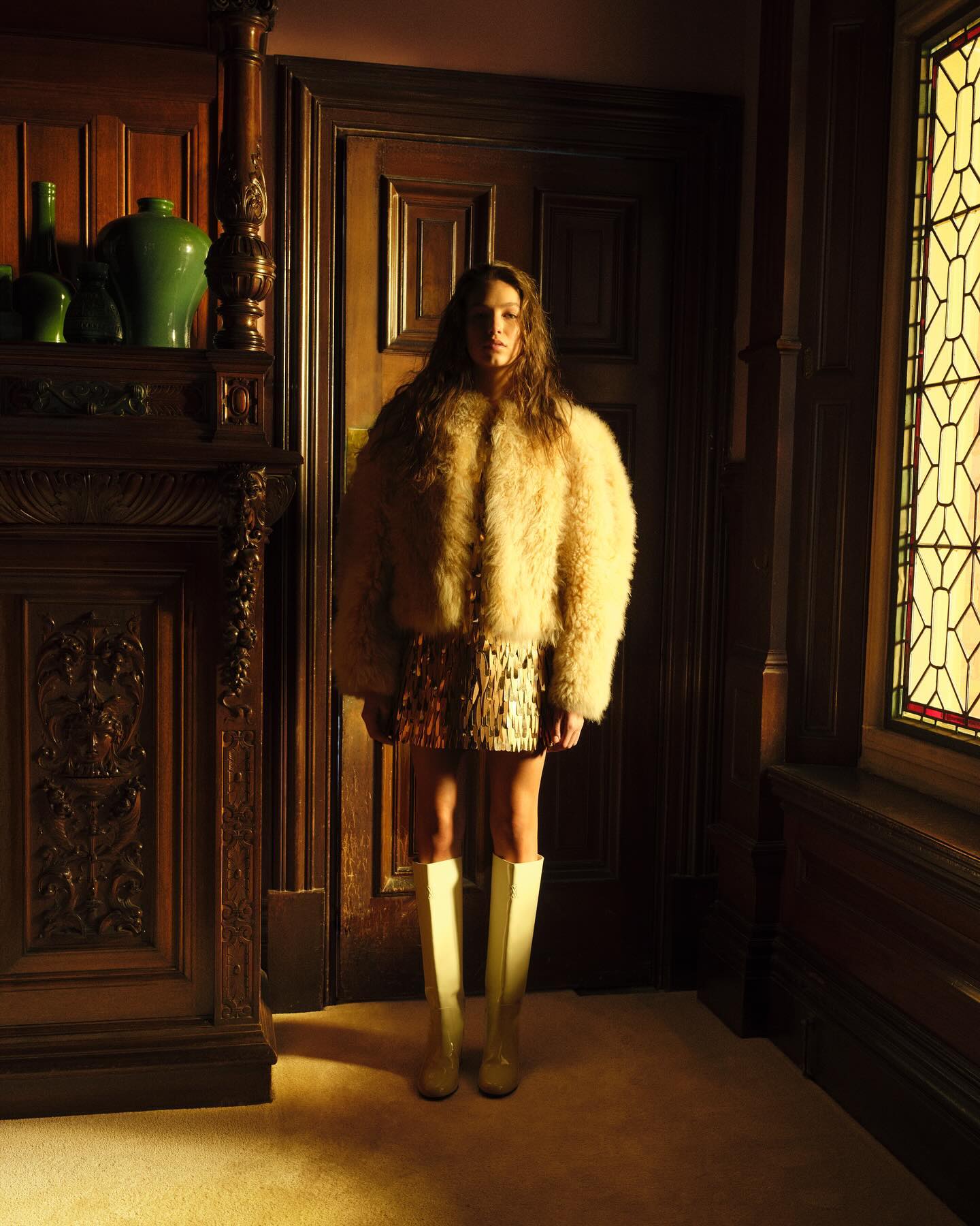 Woman in a fur coat, yellow boots, and a sequin skirt poses indoors in front of dark wood doors and a fireplace mantel.