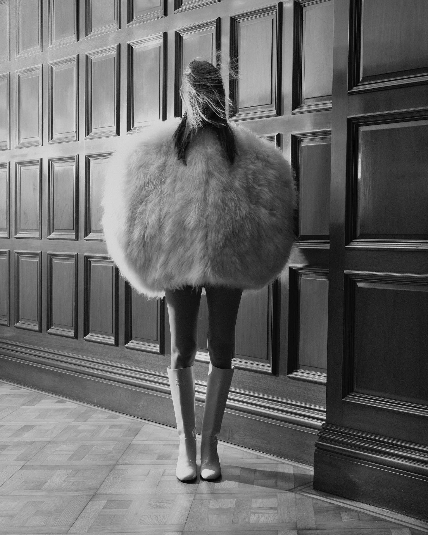 Fashion shot: Person in an oversized, round, fluffy coat, knee-high boots posing against a paneled wall and herringbone floor in black and white.
