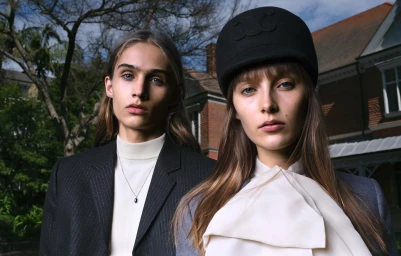 Two models outdoors wearing structured jackets, a white turtleneck, a pussy-bow blouse, and a black cap against a building exterior