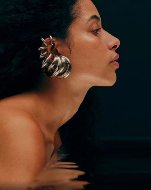 Side view of an adult woman with bare shoulders, wearing a large silver ear cuff in front of a dark background with water reflections.