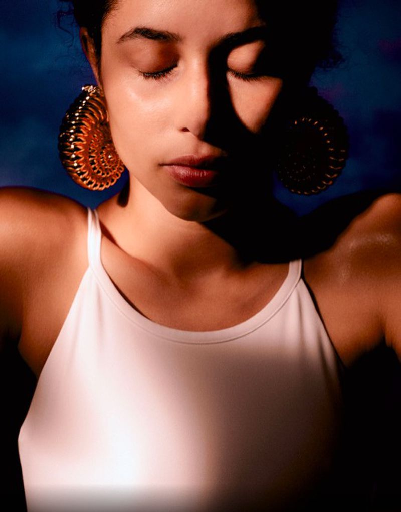 Young woman with closed eyes wears large spiral earrings. Soft light graces her face and white tank top against a dark blue background.