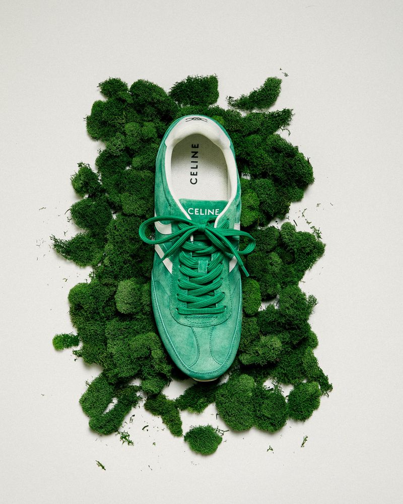 Studio shot of a green CELINE sneaker with white stripes and laces, placed on a bed of green moss, against a white background.