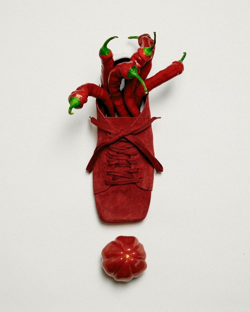 Still life with red chilies bursting from a laced red shoe, alongside a ripe tomato, creating a visually striking food composition.