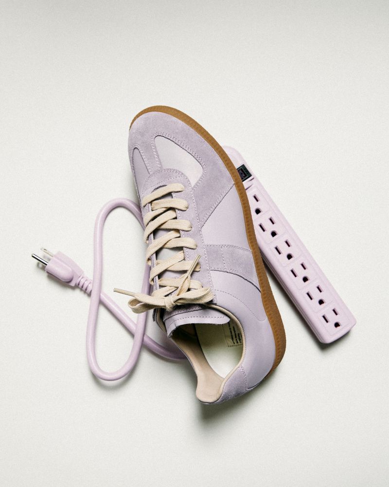 Still life studio shot of a light purple sneaker with tan trim, a matching power strip, and an integrated plug and cord on white.