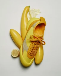 Yellow sneaker arranged vertically alongside fresh yellow squash, a halved lemon, and a mesh net on a white background.