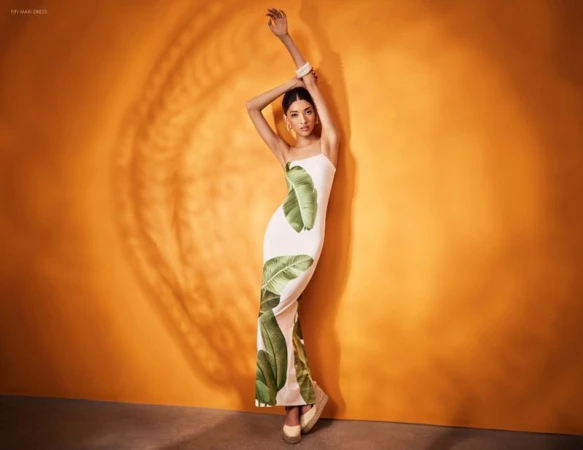 A young woman in a white dress with leaf patterns poses against an orange backdrop with light and shadow for a fashion layout.