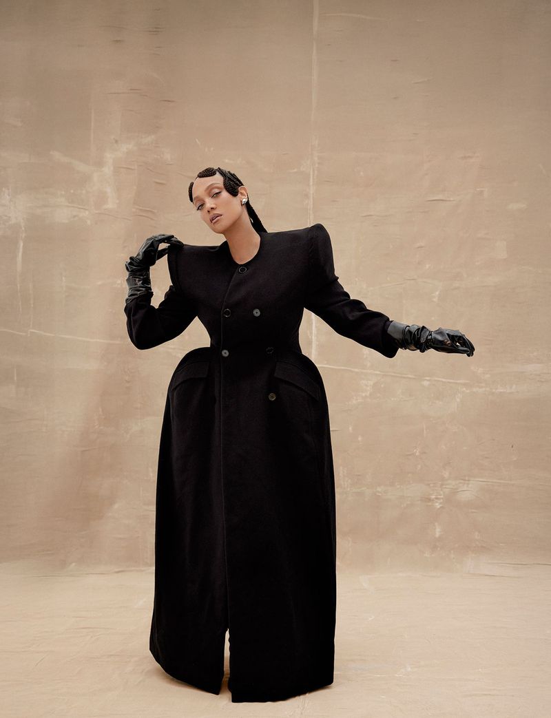 Fashion shot of a stylish woman in a long black coat and gloves striking a pose in front of a tan backdrop, fashion editorial.