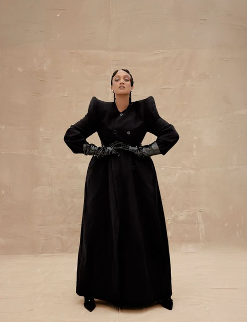 Fashionable woman posing in a black coat, gloves, and heels against a neutral backdrop, showcasing editorial style and elegant outerwear.