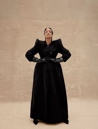 Fashionable woman posing in a black coat, gloves, and heels against a neutral backdrop, showcasing editorial style and elegant outerwear.