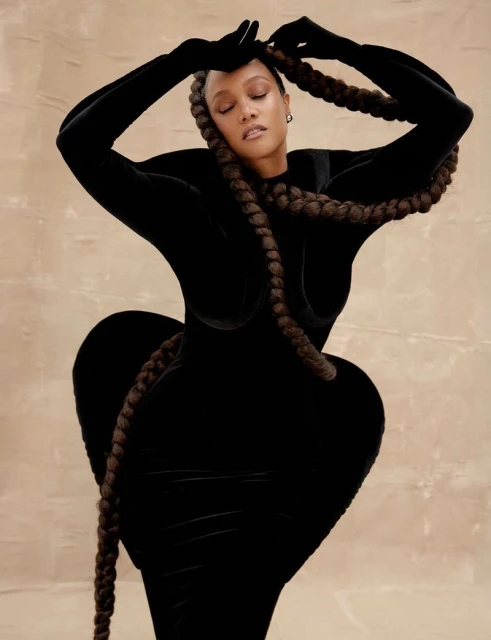 Woman in a sculptural black dress poses with long braids against a neutral background in a fashion or beauty context.