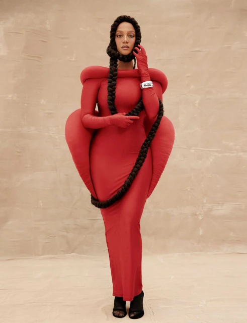 Woman poses in a red gown with braided extensions that drape around her neck and shoulders against a neutral backdrop in a studio setting.