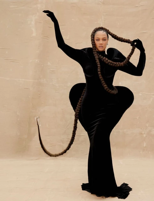 Model poses in a black velvet gown with long braids, standing against a neutral backdrop with arms raised in a studio setting.