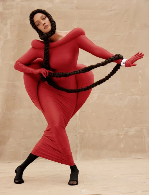 Fashion model in a draped red dress poses in a studio with a dramatic braid detail and matching gloves. Red gown has exaggerated form.