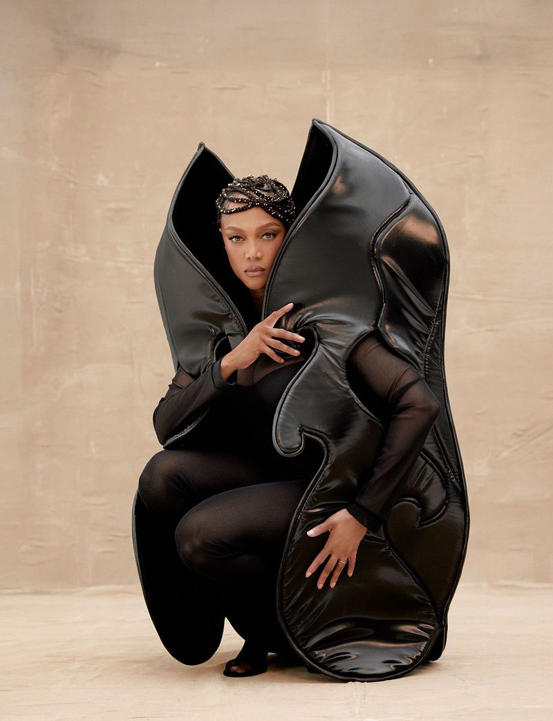 A striking image of a single female model crouching in a photo studio wearing a large, sculptural black leather garment, artistic and dramatic.