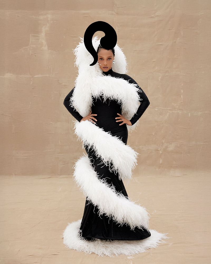 Elegant adult female in black gown with white feather boa and large black headpiece poses in a studio with neutral tan background.
