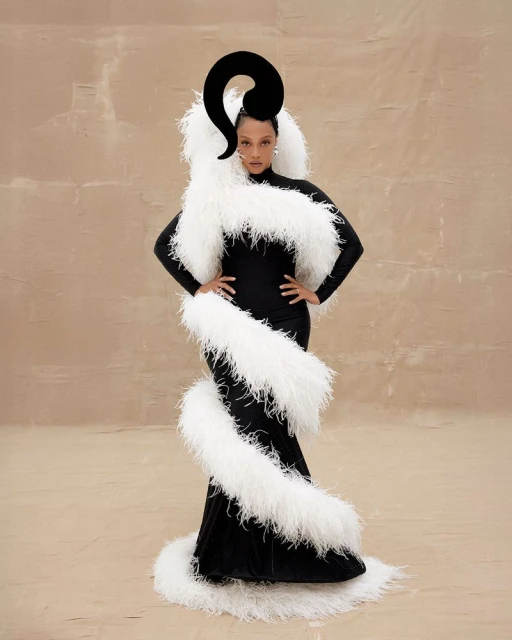 Elegant adult female in black gown with white feather boa and large black headpiece poses in a studio with neutral tan background.