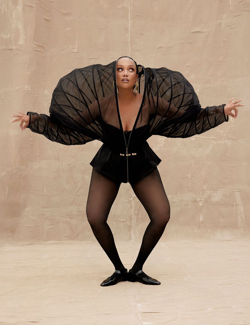 Woman in avant-garde fashion strikes a dance pose in a studio, wearing a black bodysuit, tights, and architectural tulle sleeves and headpiece.