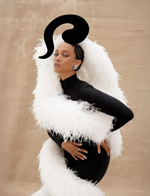 Woman posing in black bodysuit and white faux fur wrap with black sculptural headpiece against a tan textured background.