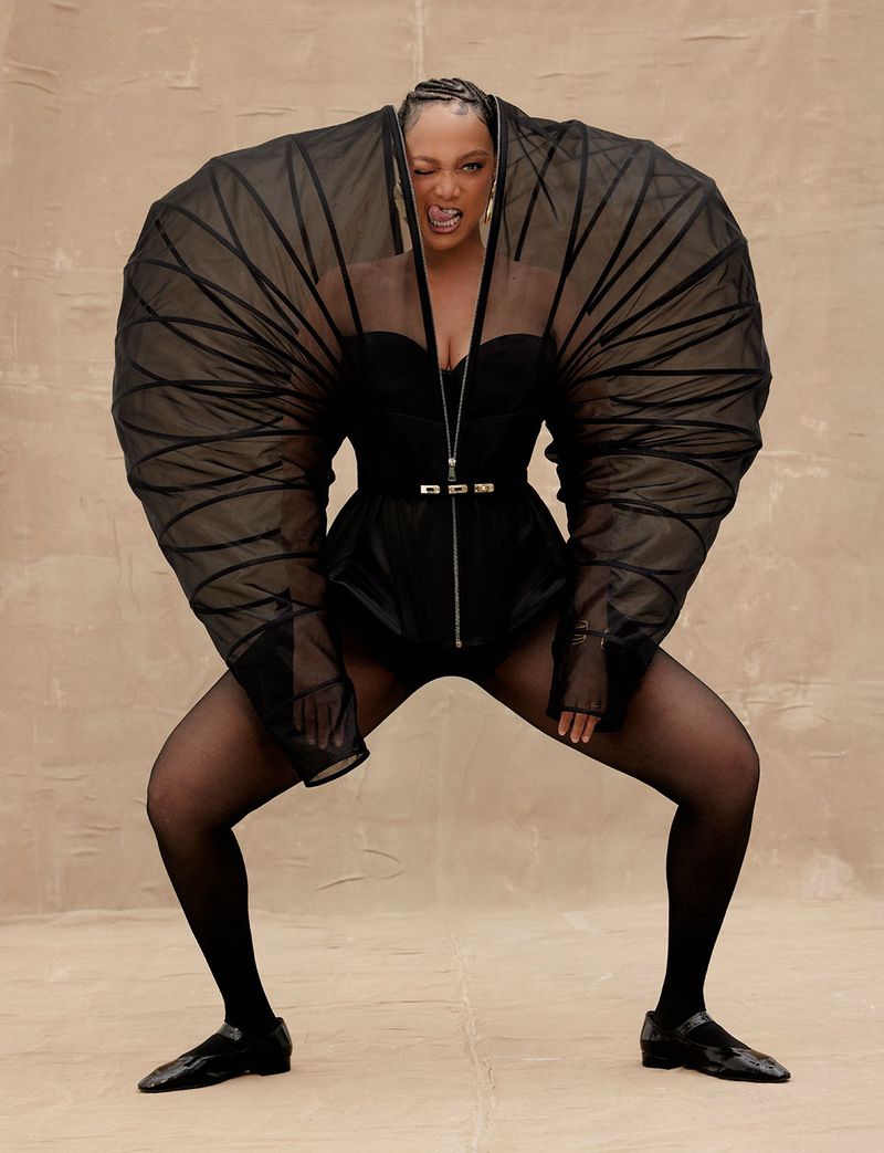 Woman in black bodysuit and tights strikes expressive pose. Black dress with tulle balloon sleeves. Neutral studio backdrop. Fashion shot.