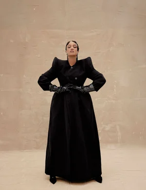 Tyra Banks in a black coat with padded shoulders and leather gloves against a neutral textured background.