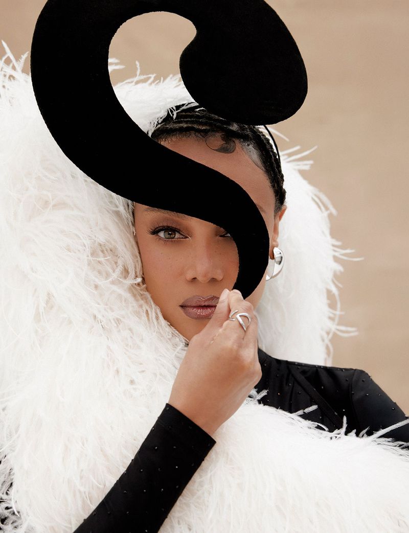 Tyra Banks holding a large black sculptural shape in front of her face, wearing a white feather boa and black top