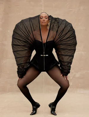 Tyra Banks posing in a structured black outfit with dramatic sheer fan-like sleeves against a neutral textured background