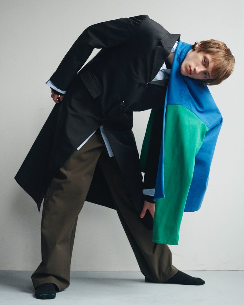 Fashion photo of a slender male adult with brown hair, wearing a long black coat and green pants, leaning with a scarf indoors.