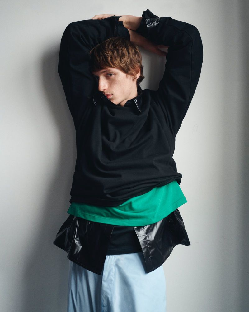 Young adult fashion model poses indoors in layered clothing, including a black top, green shirt, and shiny black jacket against a white wall.