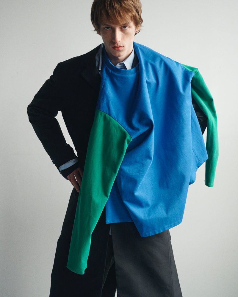 Fashion shot of an adult male posing indoors wearing a blazer over a light blue shirt, holding a color-blocked shirt.