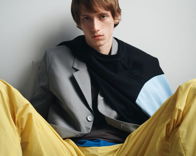 Model wearing a grey blazer, black pullover, and yellow trousers against a white seamless background