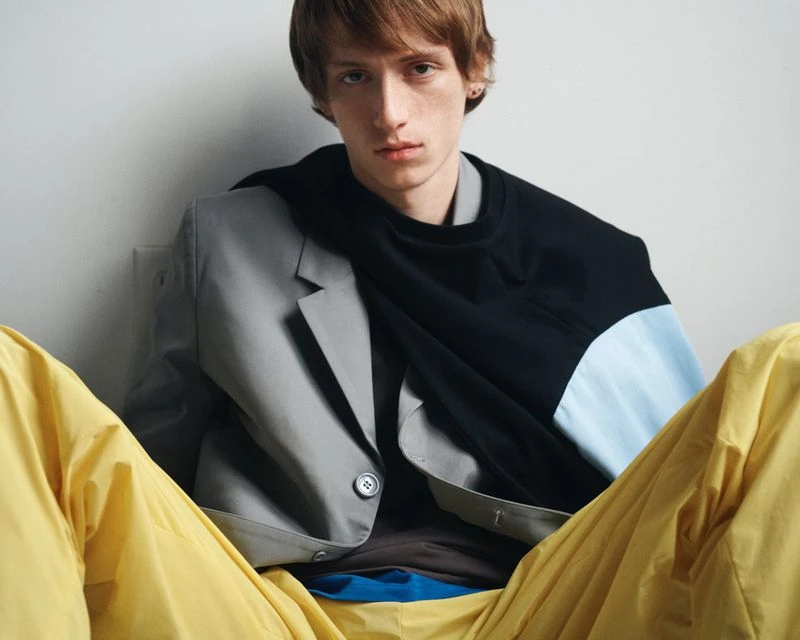 Model wearing a grey blazer, black pullover, and yellow trousers against a white seamless background