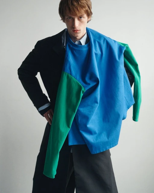 Young male model wearing layered colorful long-sleeve top over black coat against a plain white studio background.