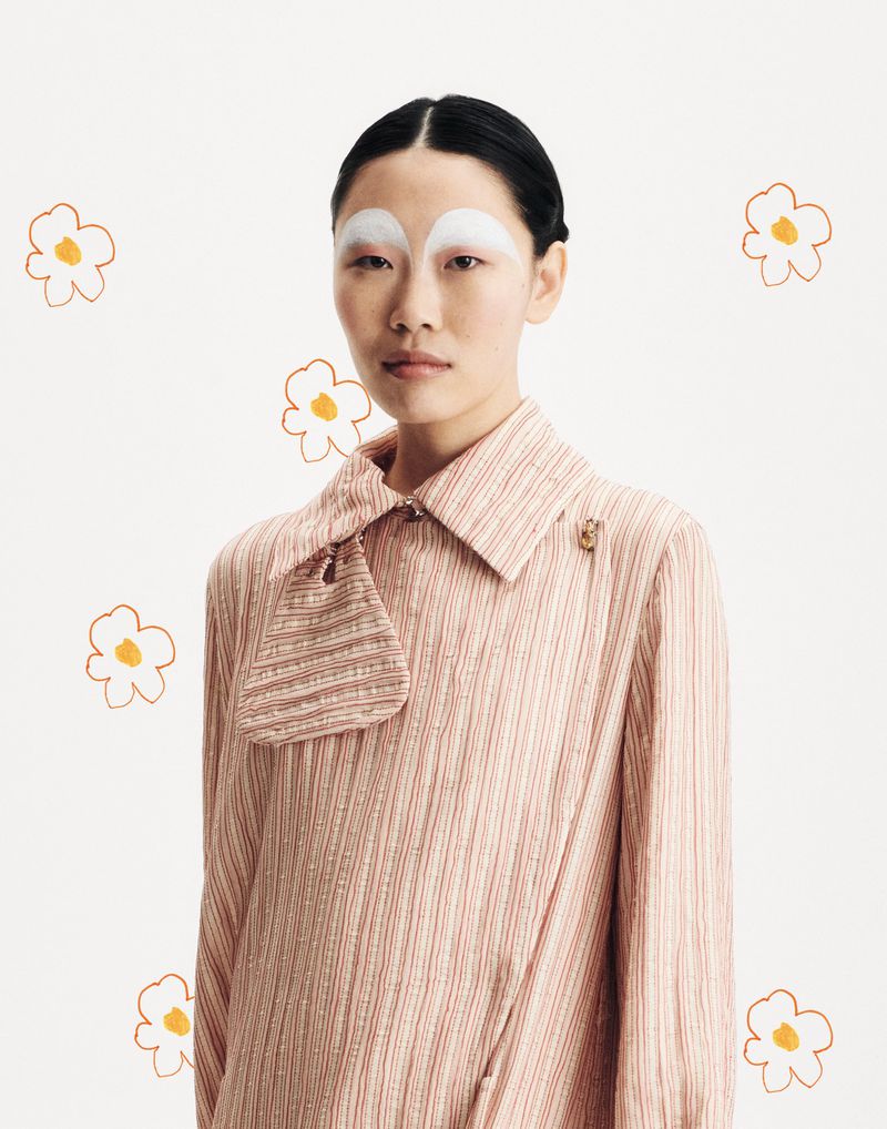 Portrait of an adult female with painted white eyebrows and a striped collared shirt, set against a backdrop of flower illustrations.