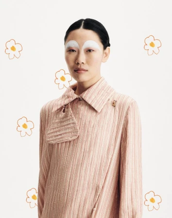Portrait of an adult female with painted white eyebrows and a striped collared shirt, set against a backdrop of flower illustrations.
